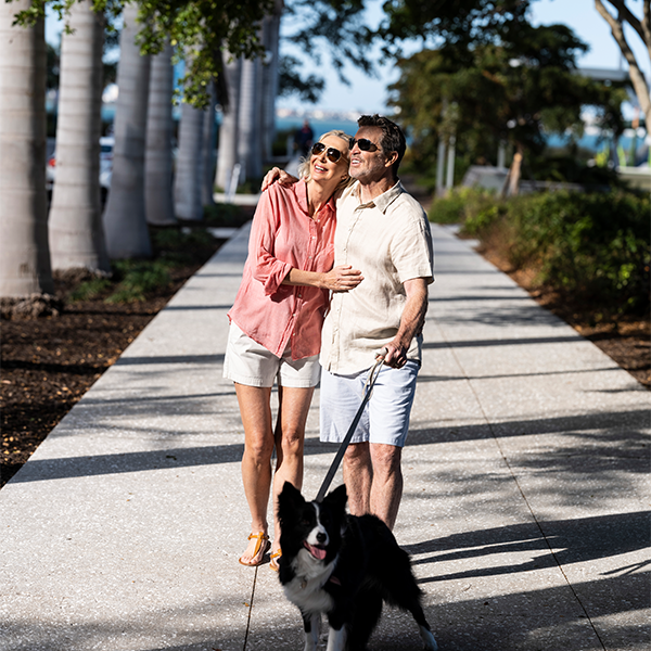 Couple walking black and white dog on shaded park path near waterfront.