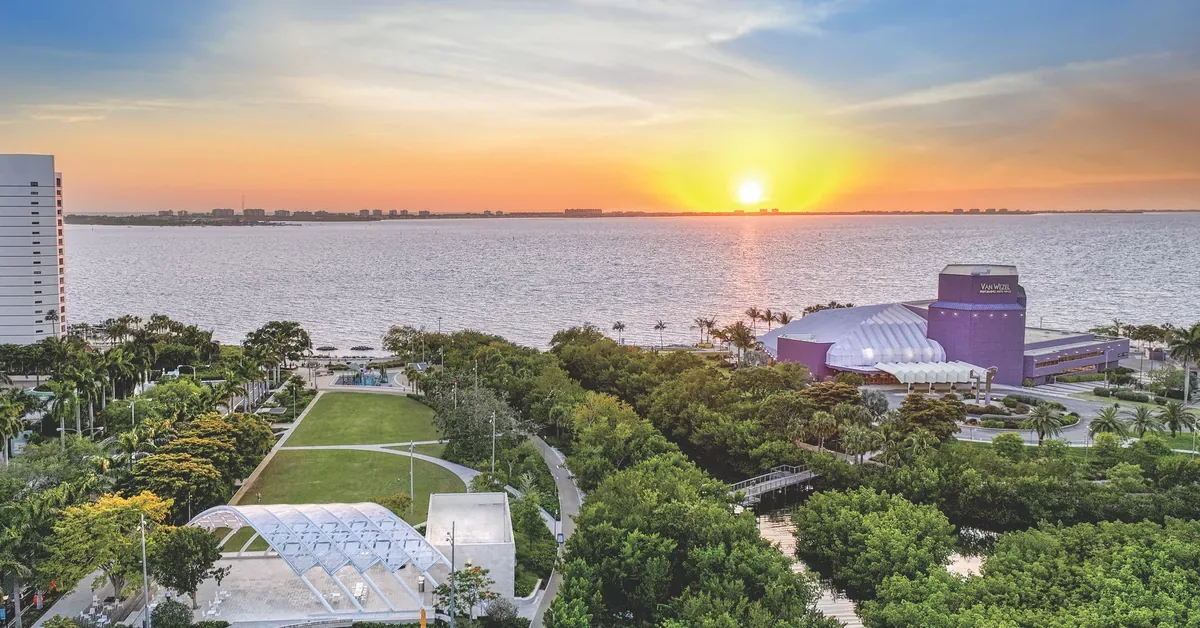 Stunning aerial view of waterfront park with amphitheater, lush greenery, and modern buildings overlooking the bay at golden hour sunset.
